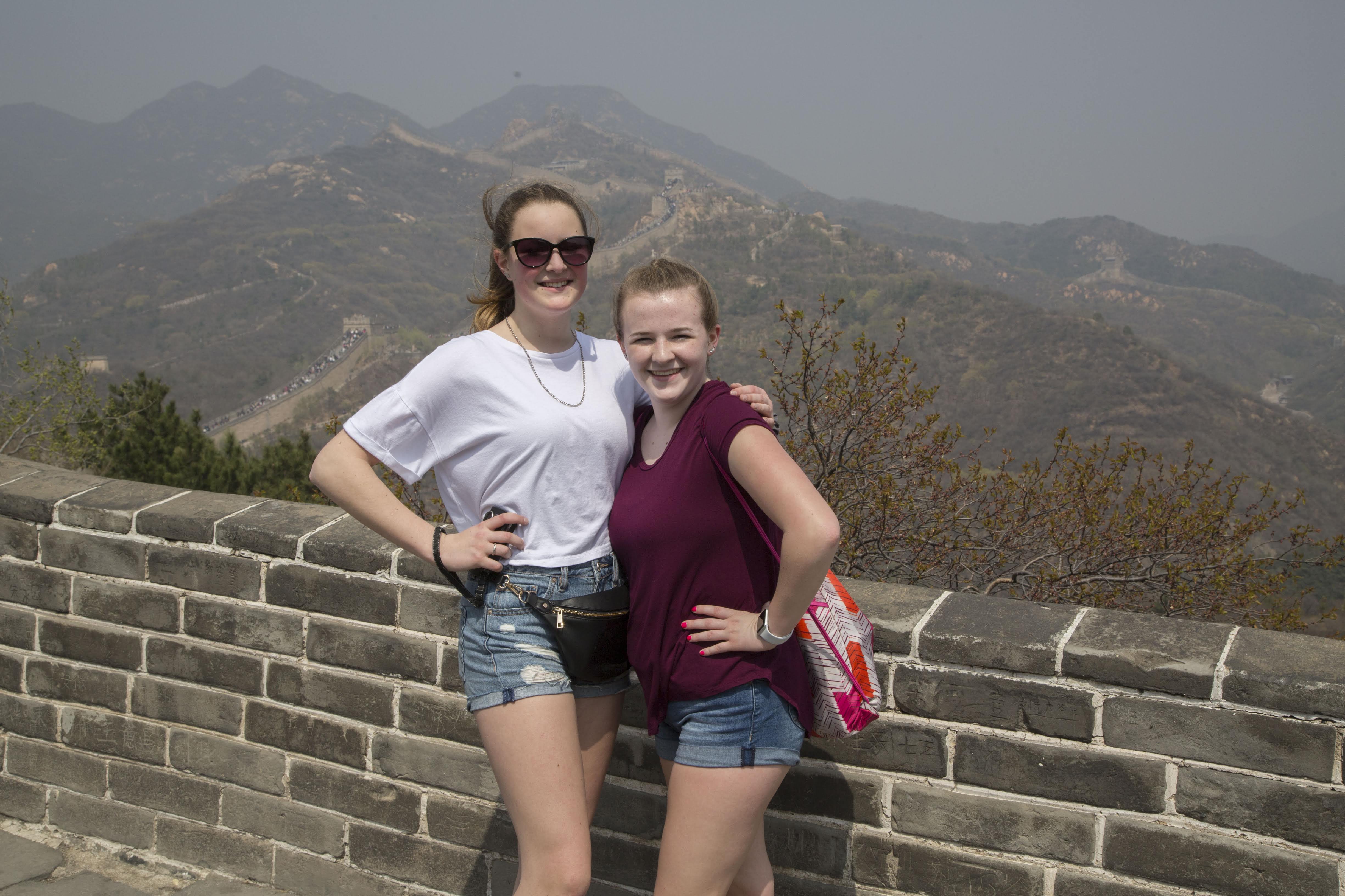 Photo of two female students atop the Great Wall of China with the mountains behind them.